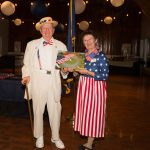 Billie and Robert at the 2019 Chapter and Partner Conference dressed in patriotic themed outfits holding an overview image of Freedoms Foundation Valley Forge campus and the American flag