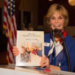 Sandy Cook of the Sacramento Chapter stands at a podium and holds a scrap book of her families 2018 trip to Valley Forge, Philadelphia and New York City.