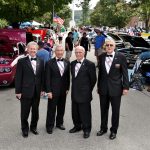 Four adults in black suits standing in front of cars