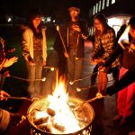 Group of students huddle around a fire roasting marshmellows
